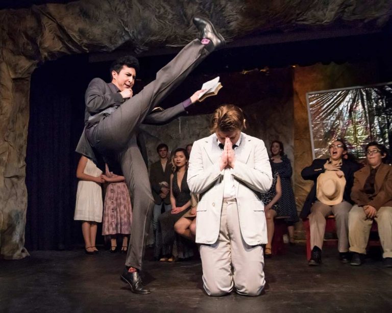 Theater performance showing actor in gray suit doing high kick over kneeling actor in white suit on stage