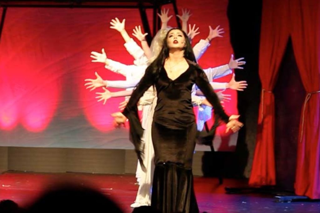 Woman in black dress performing on stage with backup dancers raising arms behind red curtains in theater production