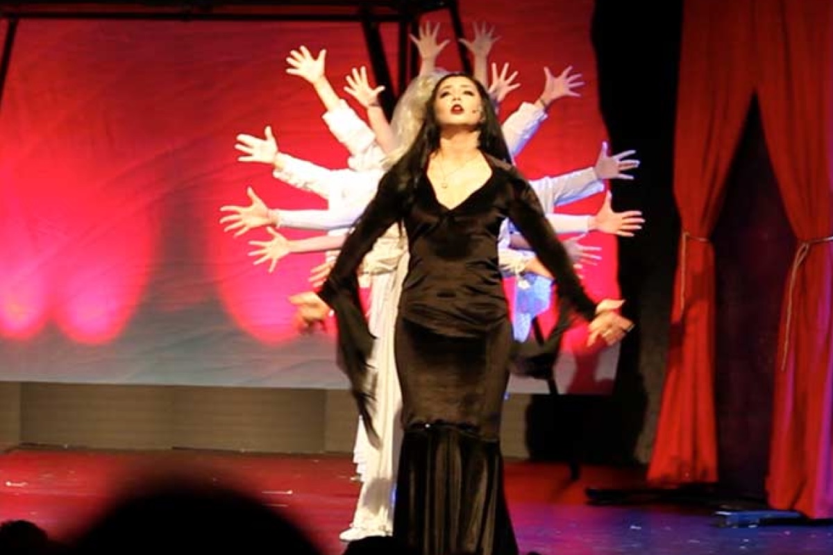 Woman in black dress performing on stage with backup dancers raising arms behind red curtains in theater production