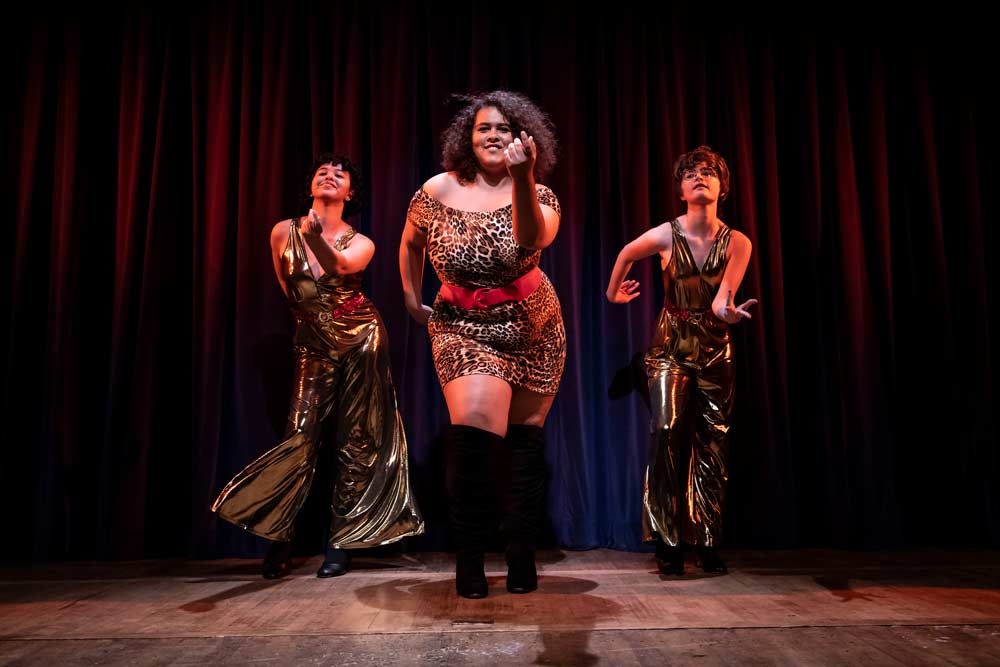 Three performers in gold and leopard print costumes dancing on stage with red curtain backdrop in theatrical production