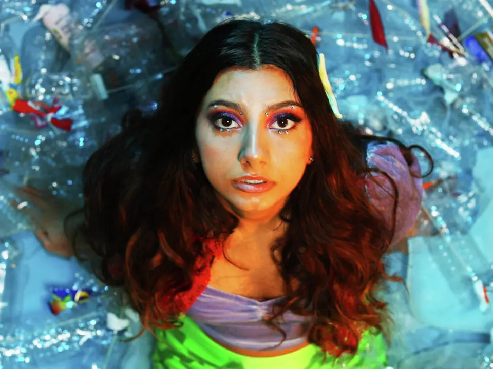 Young woman with long wavy hair and colorful makeup surrounded by shimmering blue water and floating plastic waste