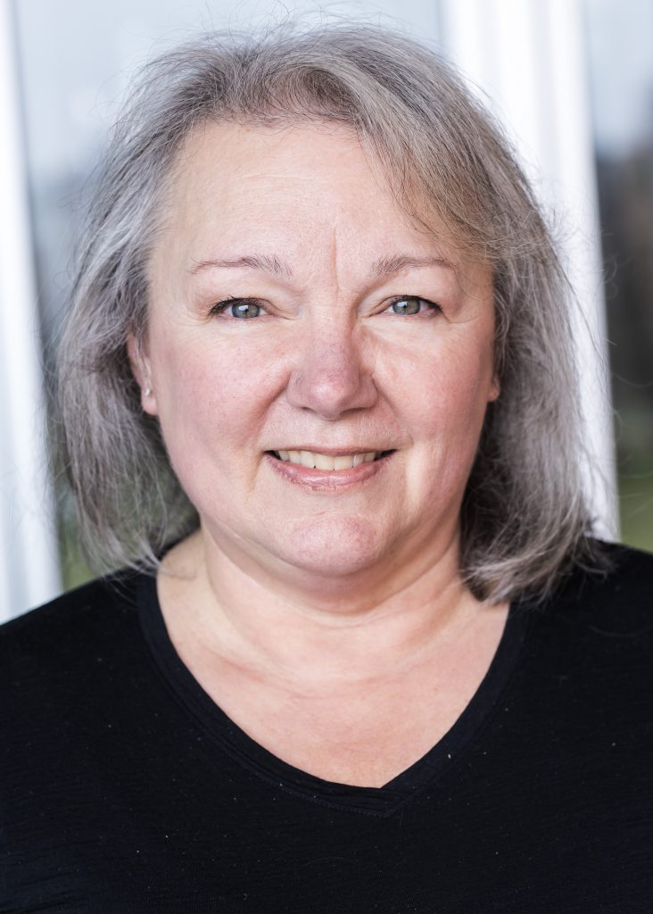 Professional headshot of smiling middle-aged woman with gray hair wearing black top against blurred office background