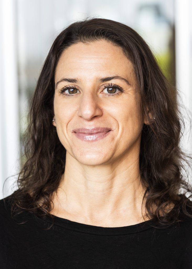 Woman with dark wavy hair and brown eyes smiling warmly at camera wearing black top in bright indoor setting