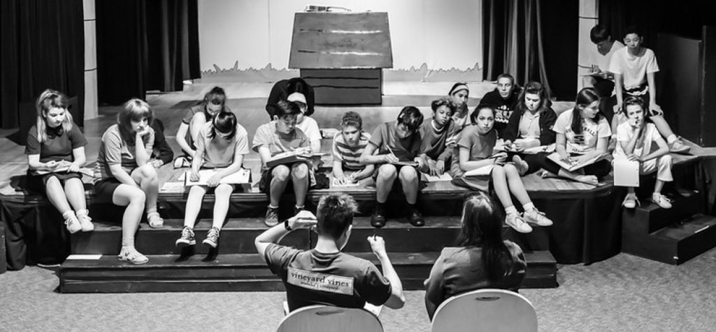 Students rehearsing on stage for school theater production, sitting with scripts in black and white photo