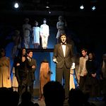 Theater performance with actors on stage in period costumes under dramatic lighting with audience silhouettes visible