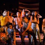 Group of diverse young people sitting together on colorful striped blanket outdoors at night around campfire