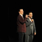 Two men in suits speaking at microphones on stage during a business presentation or conference event