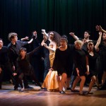 Musical theater performers in period costumes dancing on stage with dramatic lighting in ensemble production number