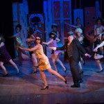 Broadway musical theater performance with dancers in colorful costumes on stage with city skyline backdrop