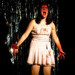 Carrie - Woman in white pleated dress with red hair standing against dark metallic backdrop for fashion photo shoot