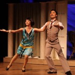 Two actors performing on stage in period costumes, holding hands while dancing or moving together in a theatrical scene.