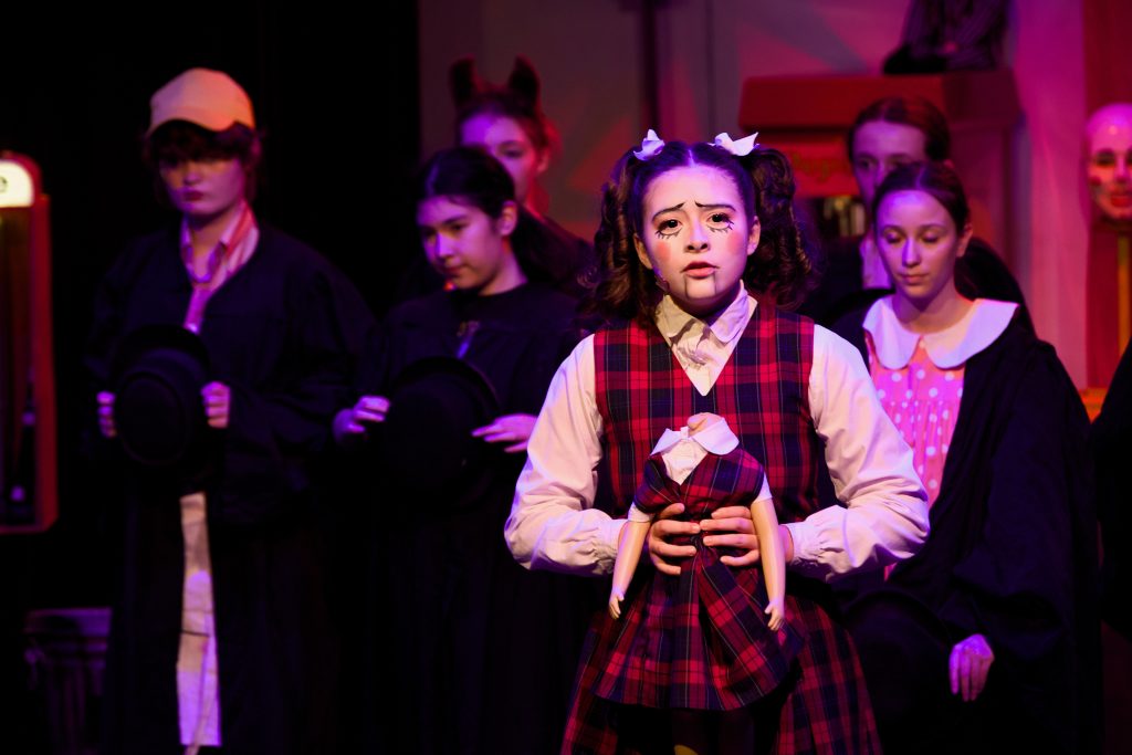 Young actress in plaid dress and pigtails performs on stage with ensemble cast in theatrical production under purple lighting.