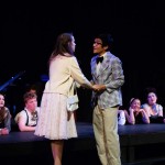 Two performers in period costumes hold hands center stage during a theatrical production with ensemble cast in background